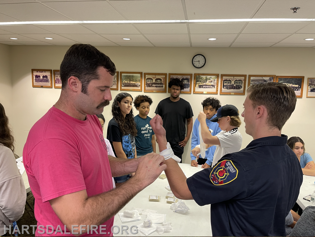 A firefighter demonstrates bandaging techniques to a group of teenagers in a classroom setting.