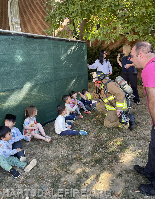 A firefighter interacts with young children sitting on the ground, while adults nearby observe the scene.