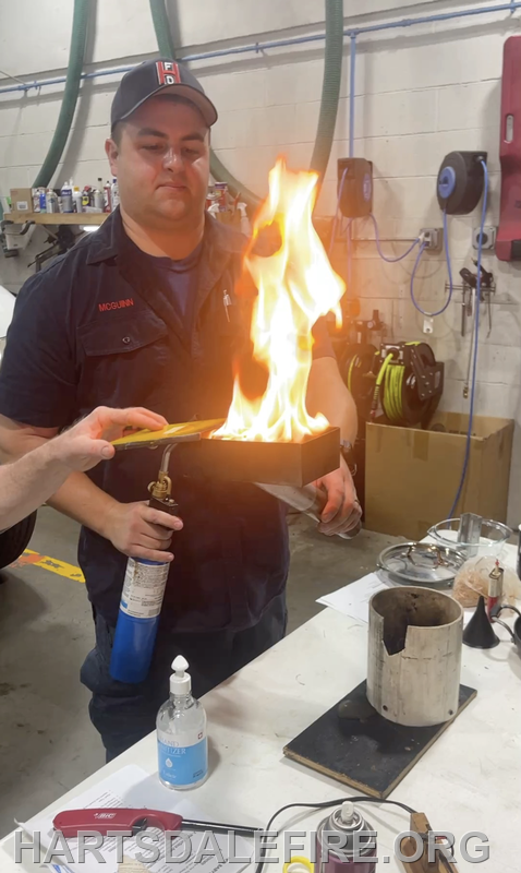 A firefighter holds a flaming box while using a torch for a demonstration, showcasing fire safety or fire behavior.