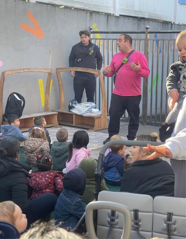 A group of children is seated on the ground while two adults, one in a pink shirt, engage with them outdoors.