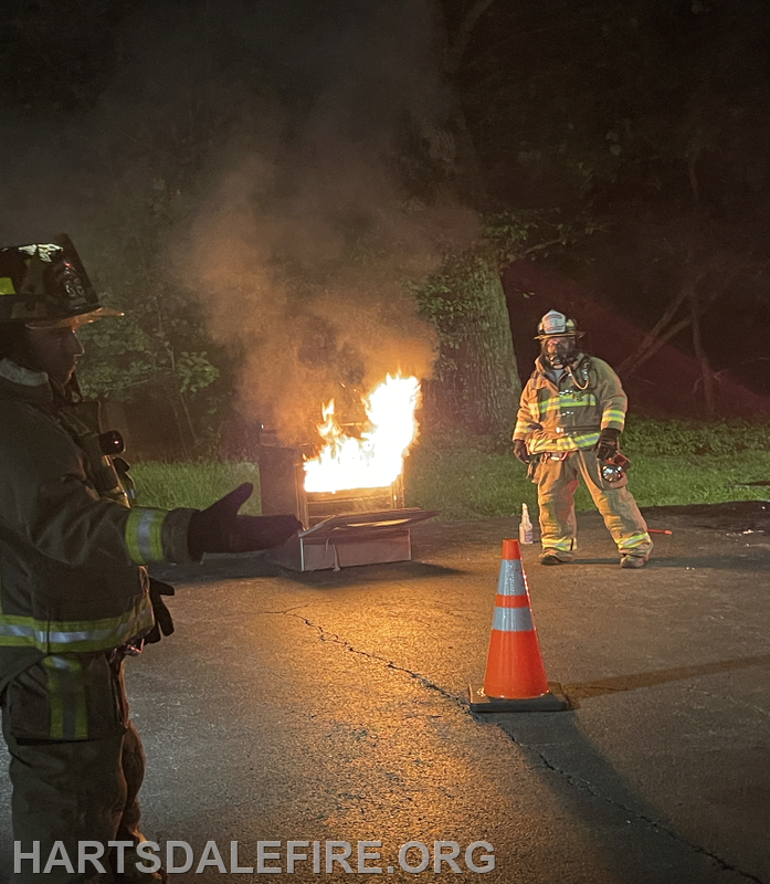 Two firefighters respond to a burning object at night, with flames and smoke visible, as they prepare to manage the situation.
