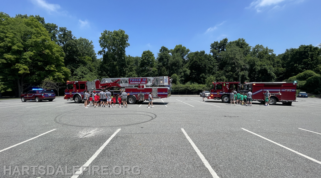 A group of children gathers around two fire trucks in a parking lot, with a police vehicle nearby on a sunny day.
