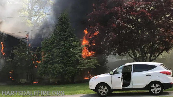 A house is on fire with visible flames and smoke. A white SUV with the door open is parked nearby.