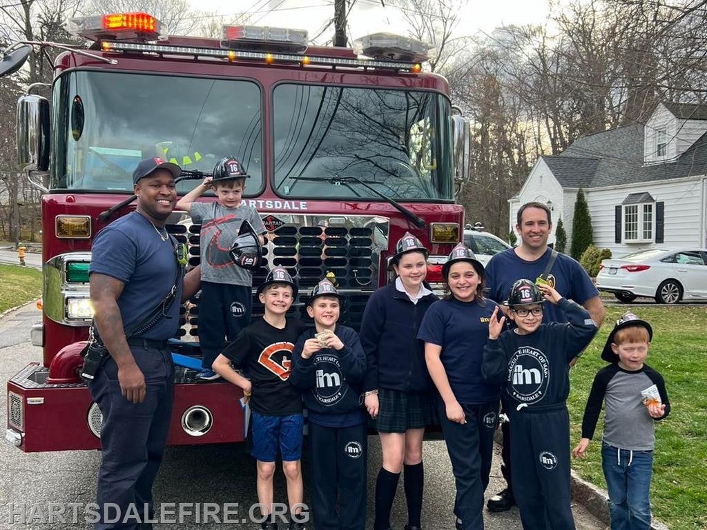 A group of kids in fire helmets poses with firefighters in front of a fire truck, smiling and enjoying the moment.