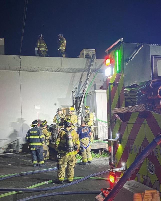 Firefighters are responding to an incident at night, with some on a rooftop and others near a fire truck and equipment.