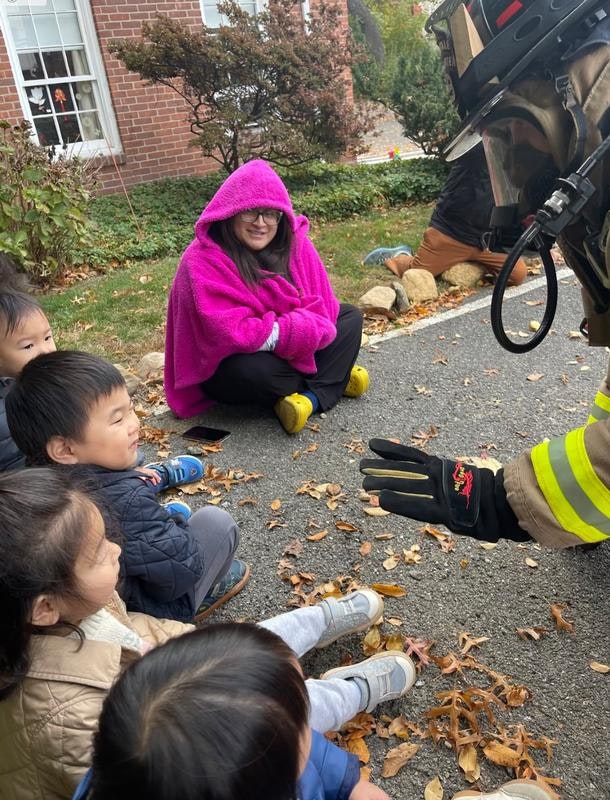 A group of children sits on the ground watching a firefighter while a woman in a pink hoodie observes from behind.