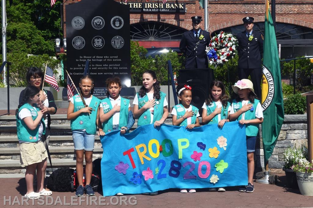 A group of girls in vests holds a "Troop 2820" banner during a memorial event; two uniformed officers stand behind with a wreath.