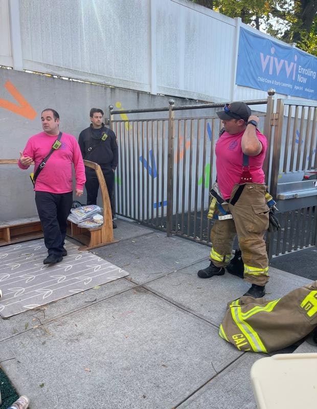 Three individuals, including two in pink shirts and one in firefighter gear, are engaging in conversation near a colorful wall.