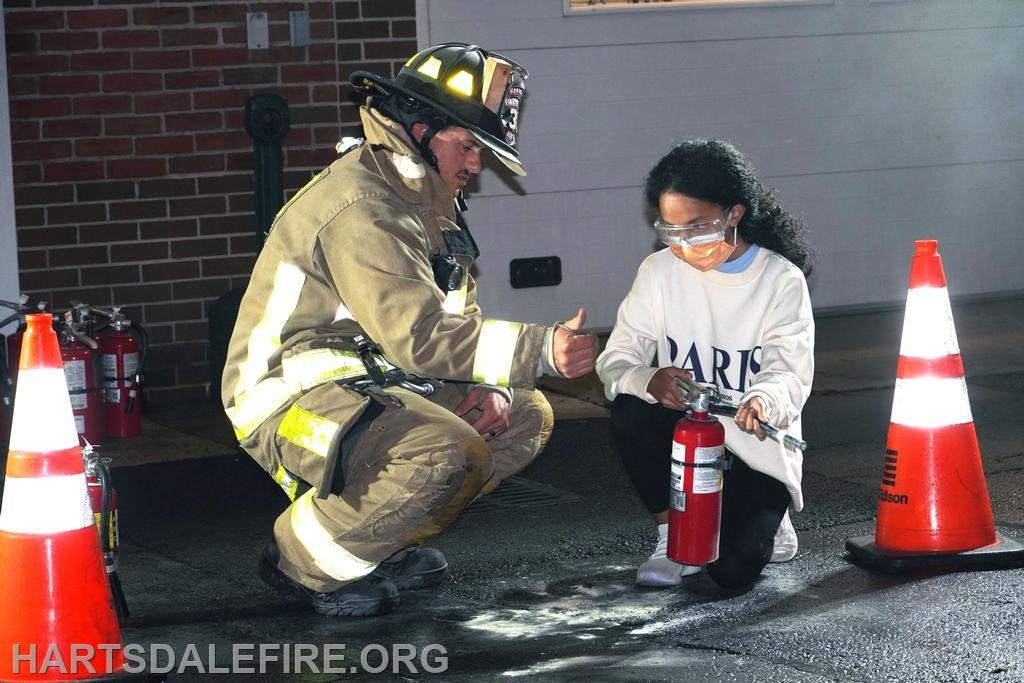 A firefighter instructs a person on using a fire extinguisher, surrounded by traffic cones.