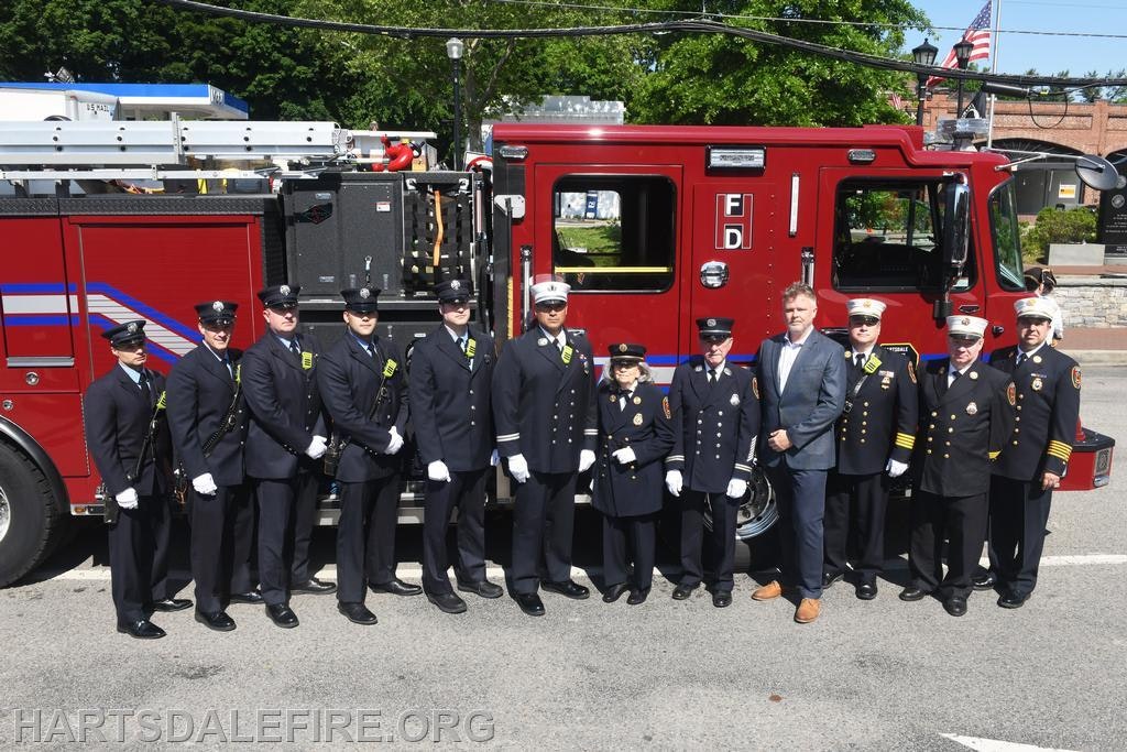 A group of uniformed firefighters and a man in a suit stand in front of a red fire truck.