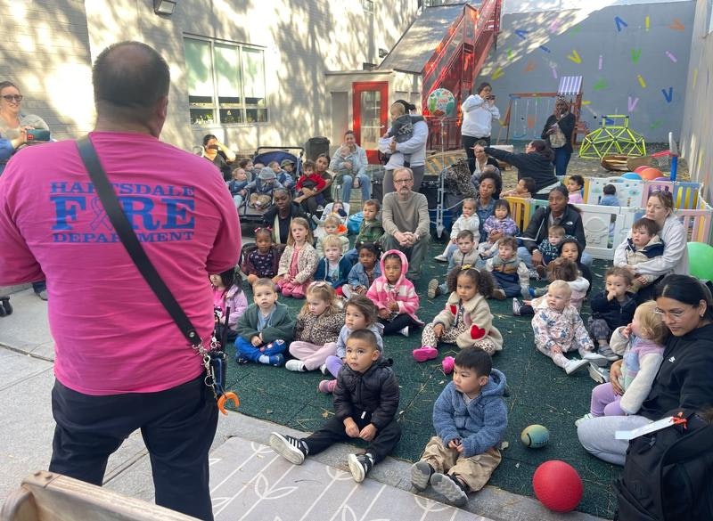 A fire department member talks to a large group of young children and adults in an outdoor setting, engaging them in learning.