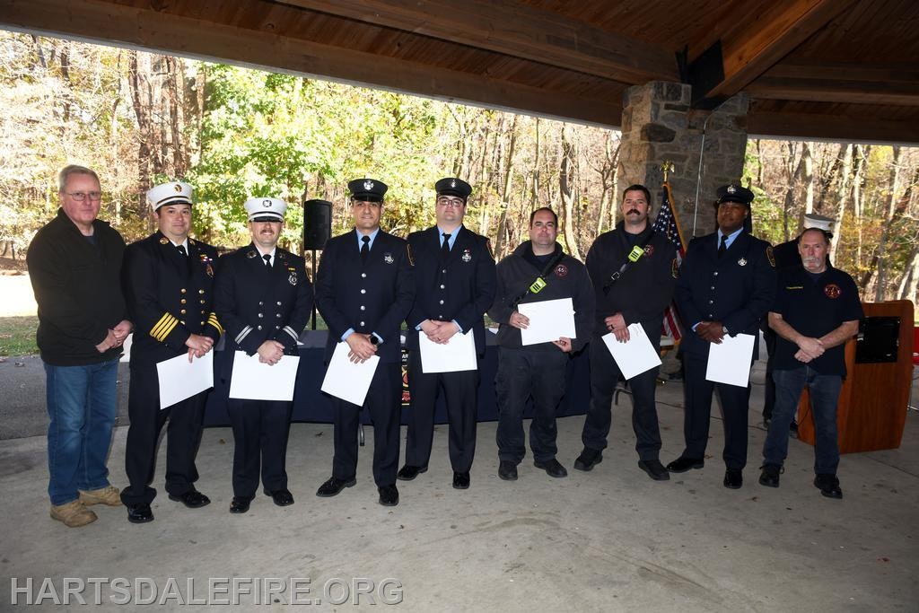 A group of firefighters and officials stands together, likely at a ceremony, holding certificates or awards outdoors under a shelter.