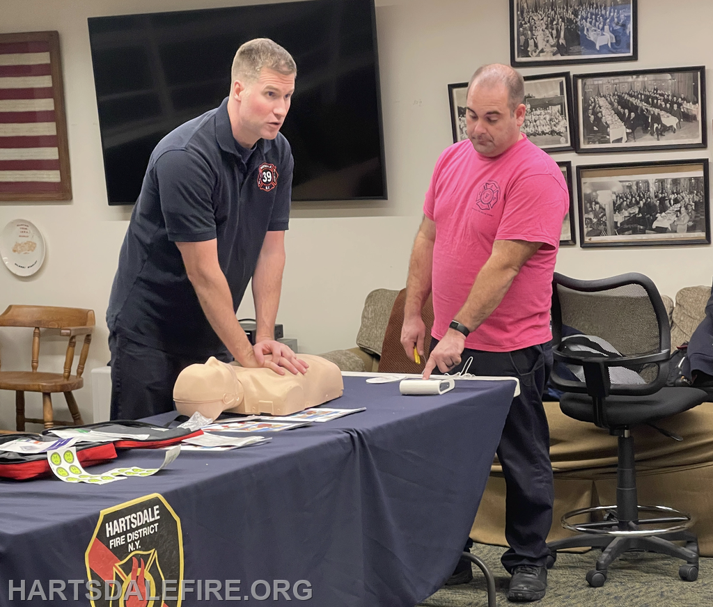 Two firefighters are conducting a CPR training session using a practice manikin on a table filled with training materials.