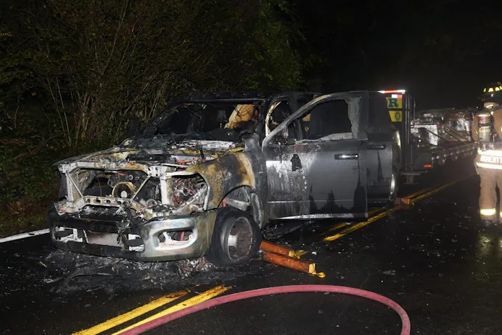 Burned-out truck on a road with firefighters nearby handling the aftermath.