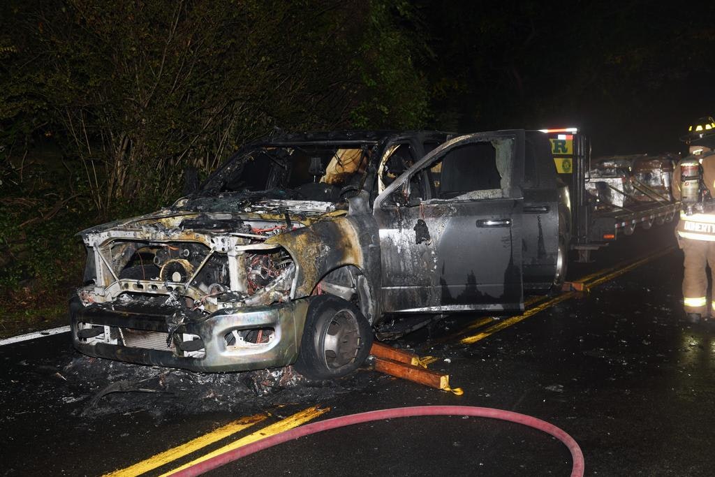 Burned-out truck on a road with firefighters nearby handling the aftermath.