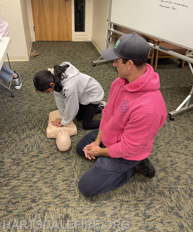 A girl practices CPR on a dummy while a man observes, in a training setting.