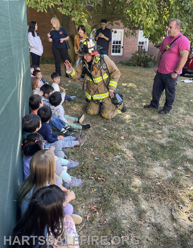A firefighter engages with a group of children outdoors, while adults watch, fostering a fun educational experience.