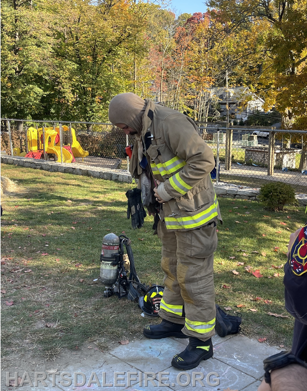 A firefighter is adjusting their gear in a park, with a playground visible in the background.