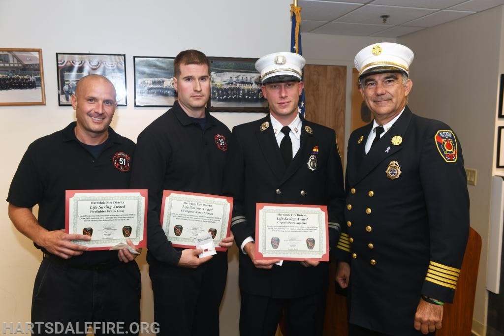 Four men in uniform holding certificates, two are in formal attire, standing in front of framed photos.