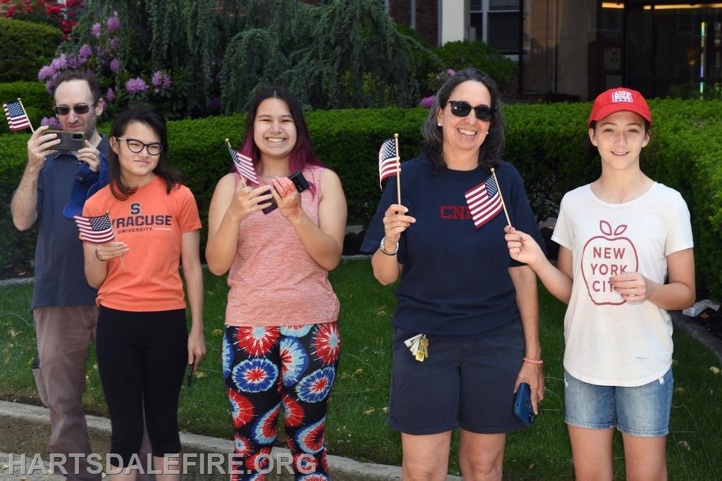 Five people holding American flags in a garden, some taking photos, wearing casual attire with various logos.