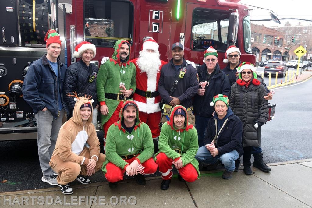 A festive group poses with Santa in front of a fire truck, dressed in holiday attire and accessories.