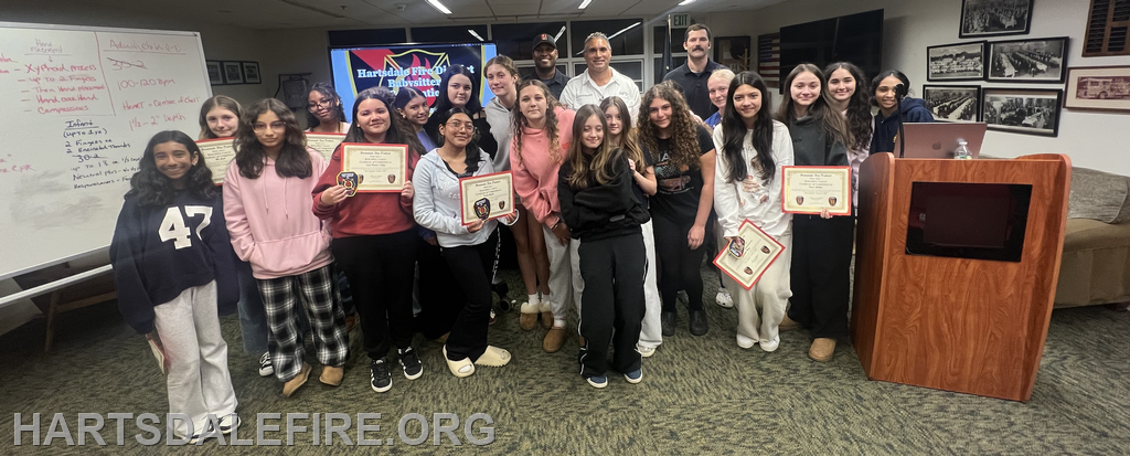 A group of girls and adults poses with awards in a room, featuring a whiteboard and a computer setup in the background.