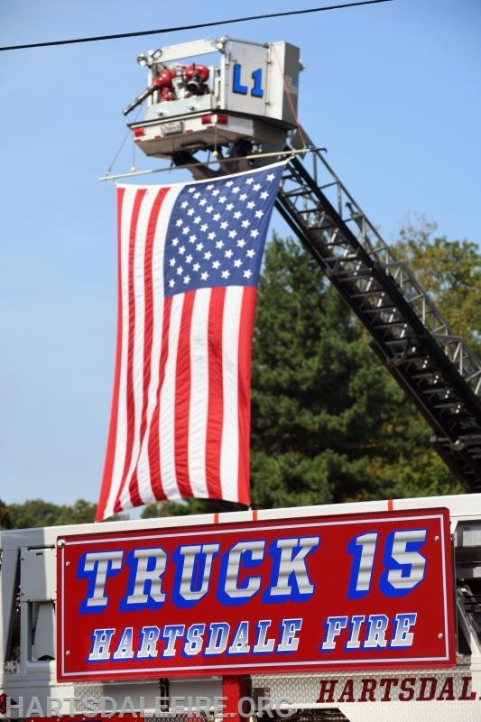 Firetruck ladder displaying an American flag, with "Truck 15" and "Hartsdale Fire" sign.