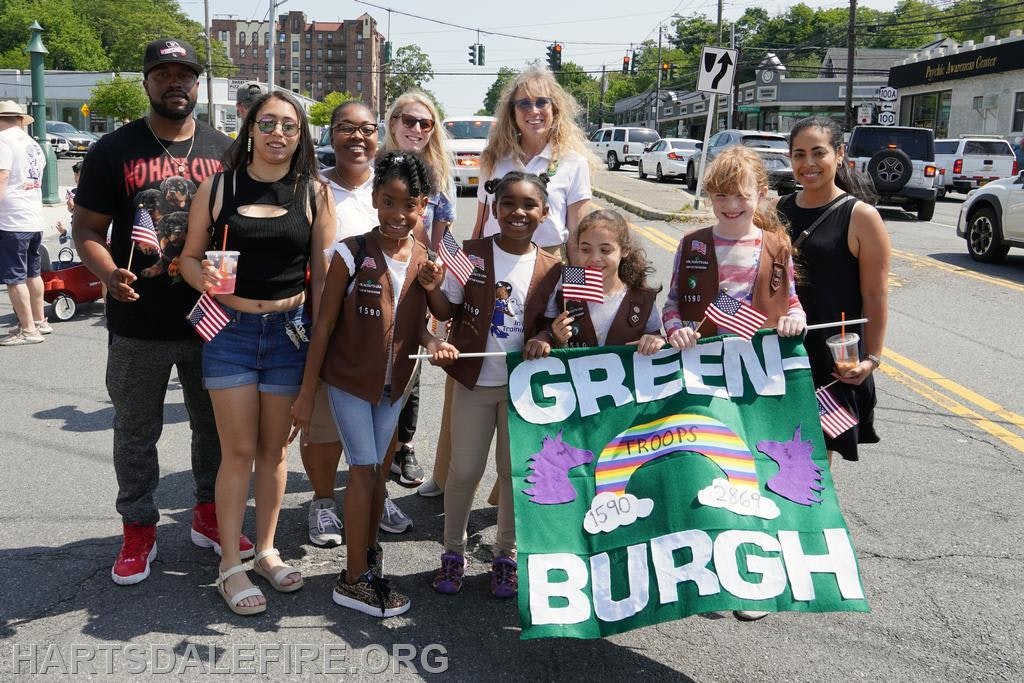 A group of people with flags, kids holding a Greenburgh troops banner, posing on a street.