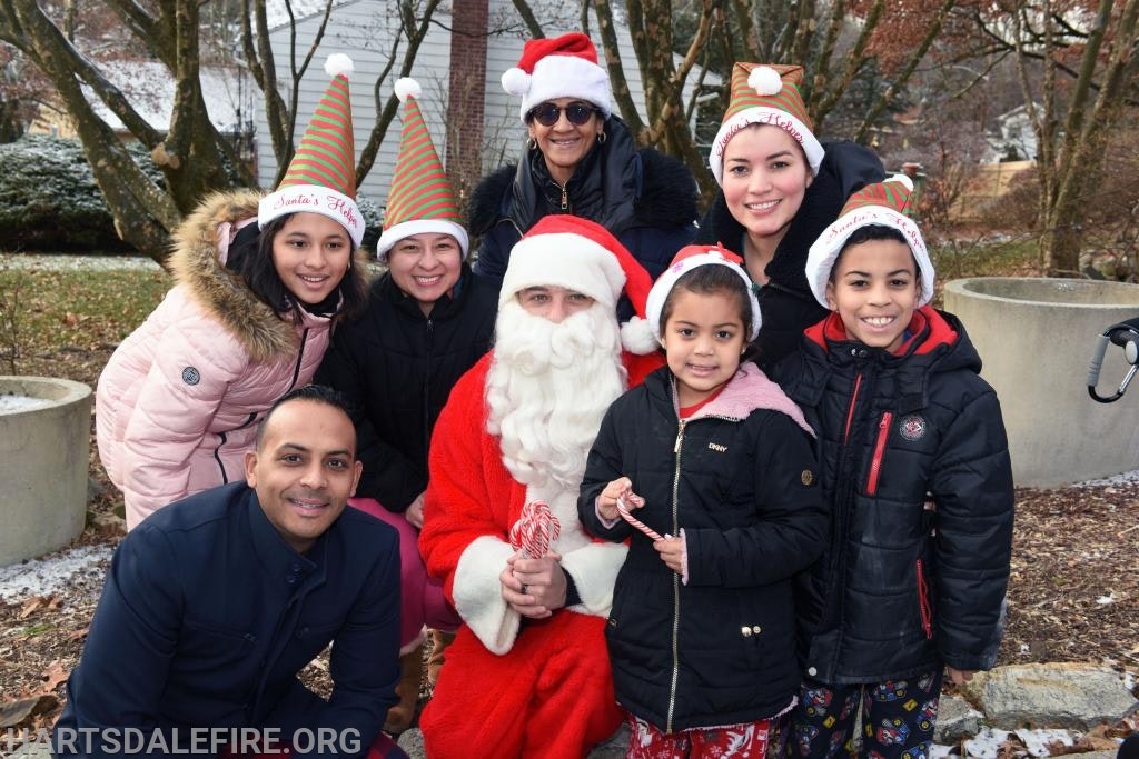 A group of people with Santa hats pose outdoors with someone dressed as Santa Claus.
