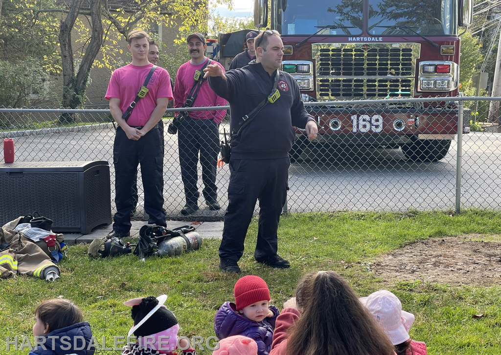 Firefighters in pink shirts interact with an audience, likely children, near a fire truck, promoting fire safety.