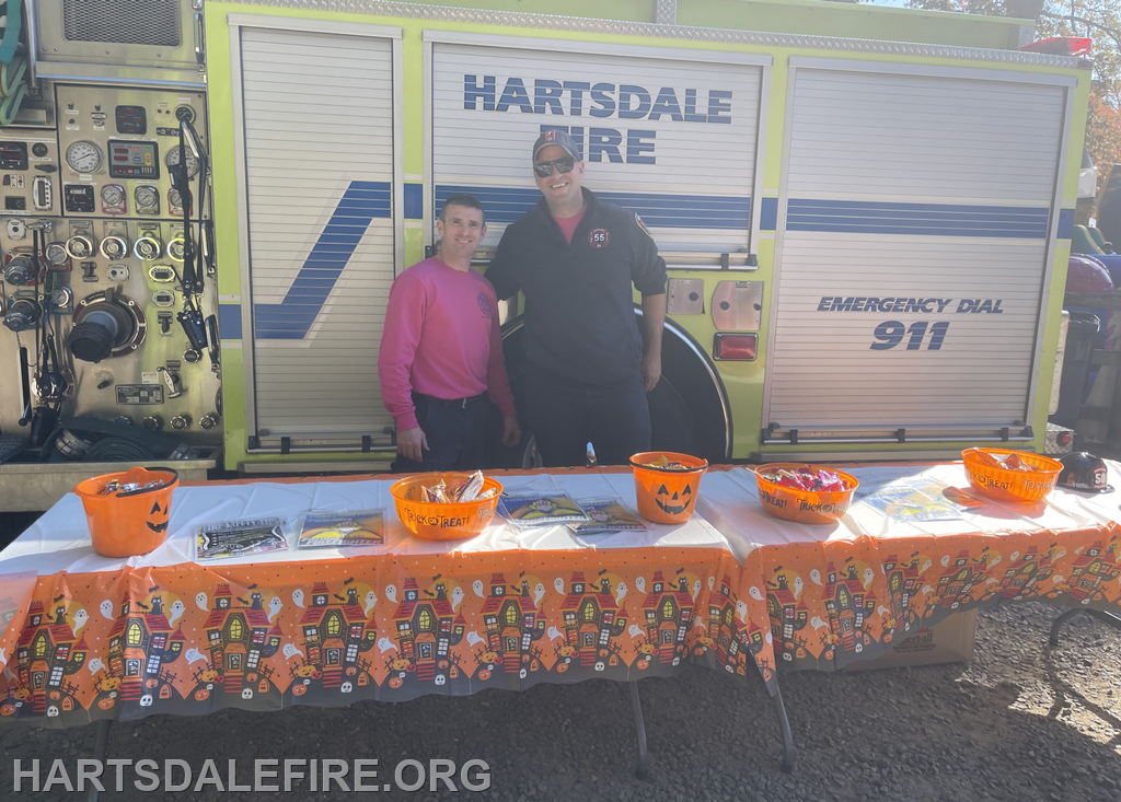 Two fire department members stand by a table with Halloween-themed decorations and candy buckets, in front of a fire truck.