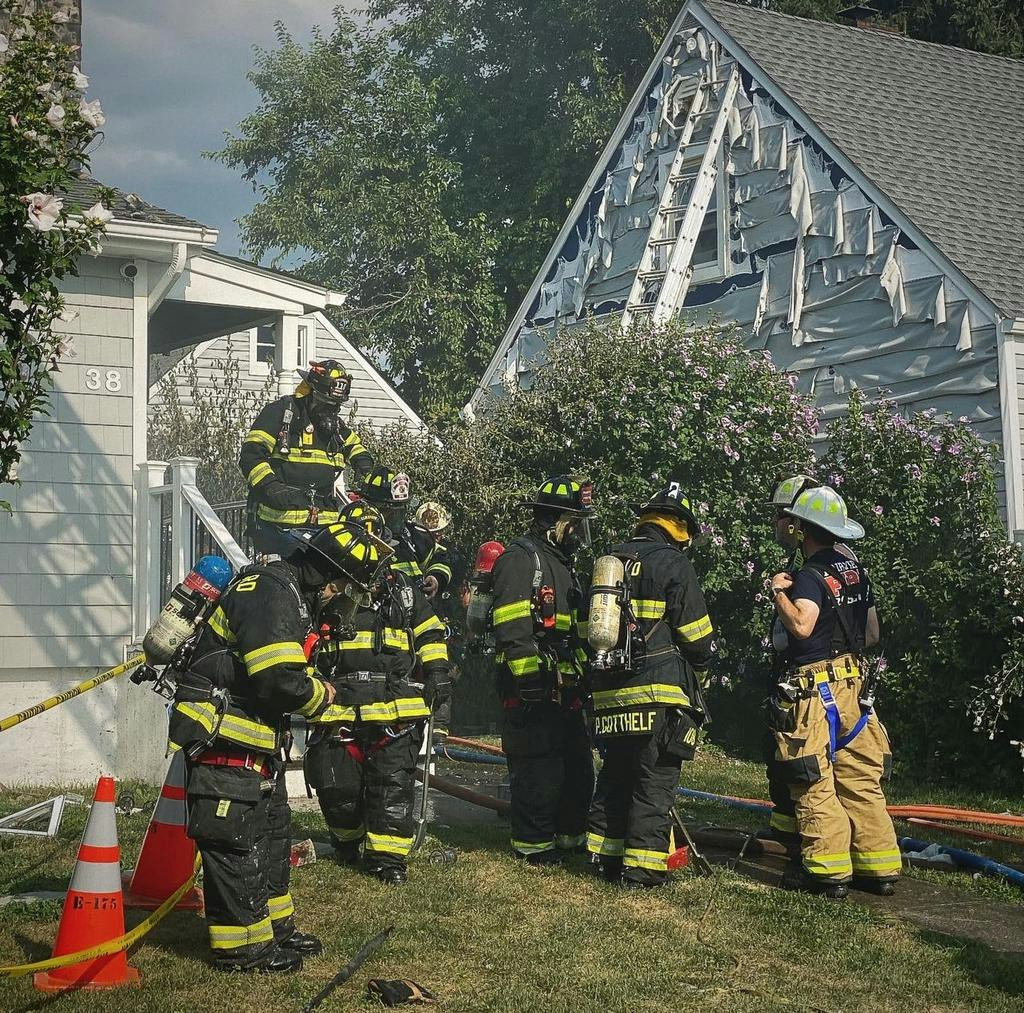 Firefighters in gear work outside a house, which has visible damage, possibly from a fire. Equipment and hoses are nearby.