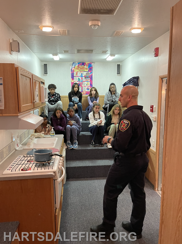 A firefighter conducts a safety lesson in a model kitchen, with kids seated attentively on steps.