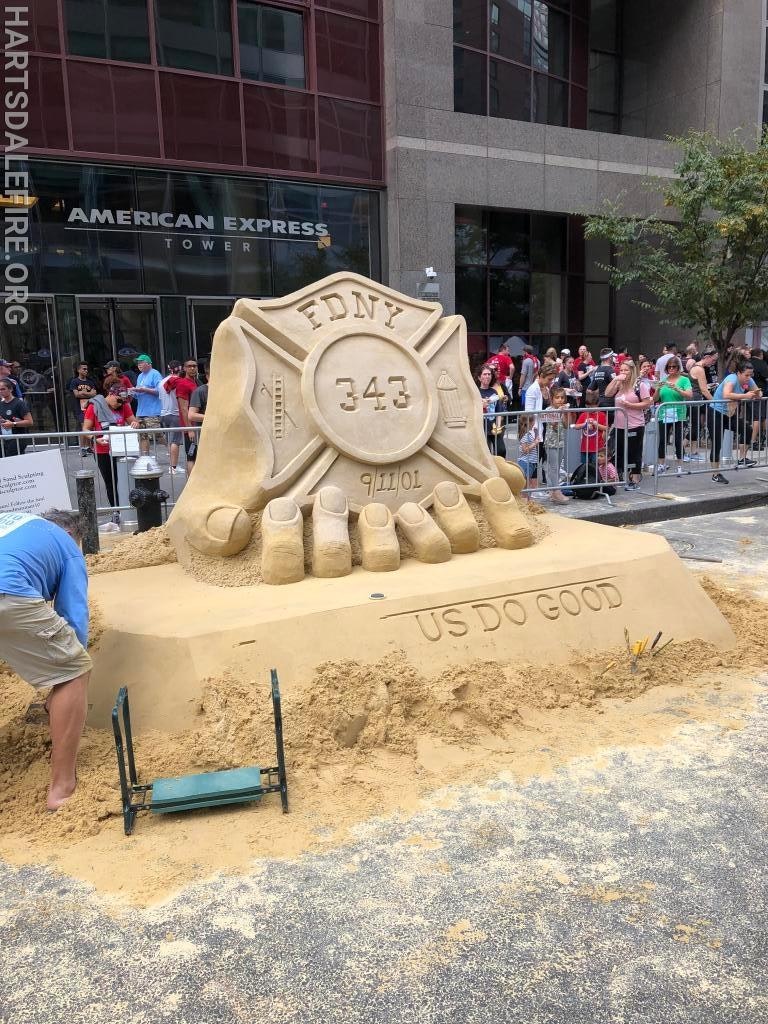 A sand sculpture of an FDNY firefighter badge with "343" and "9/11/01" carved, in front of a crowd.