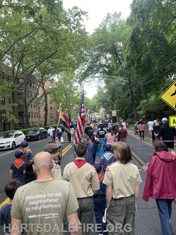 A procession of people, including scouts, walking down a street lined with trees and buildings, many carrying flags.