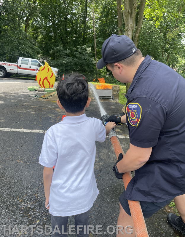 A child learns to use a fire hose with guidance from a firefighter at a demonstration near a fire truck and flames.