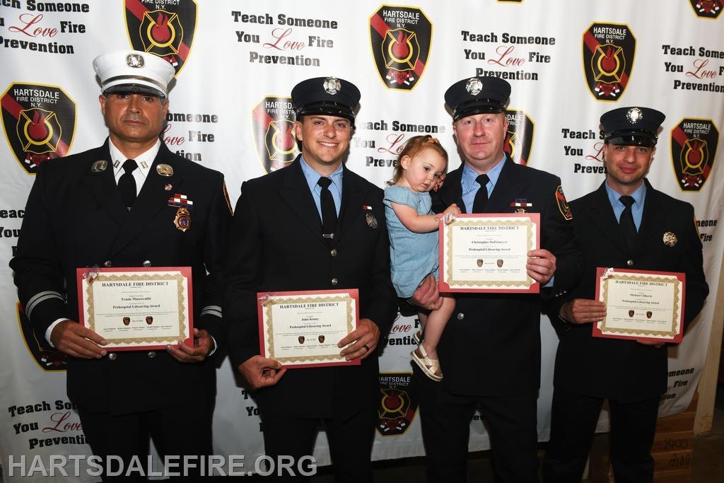 Four firefighters holding awards, one with a child, in front of a Hartsdale Fire District backdrop with fire prevention message.