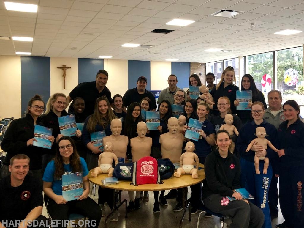 A group posing with CPR training mannequins and certification cards in a room with a crucifix on the wall.