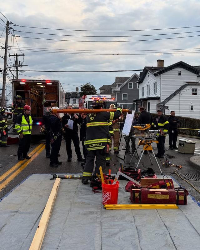 Emergency responders, including firefighters, are gathered on a street, preparing equipment and assessing a situation.