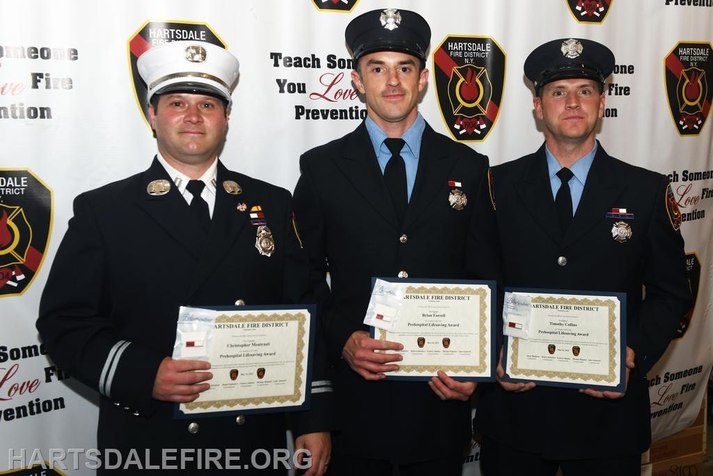 Three firefighters in uniform holding certificates, standing in front of a backdrop with fire district logos.
