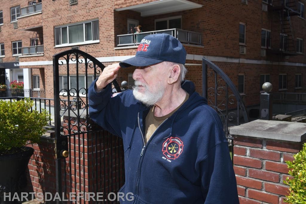 A person in a "Hartsdale Fire" cap and hoodie saluting beside a gated brick building.