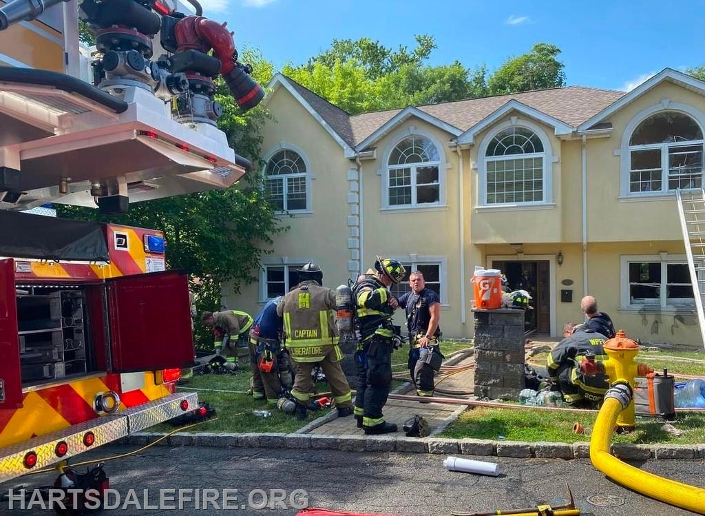 Firefighters and equipment outside a house; fire truck, hoses, and ladder visible.