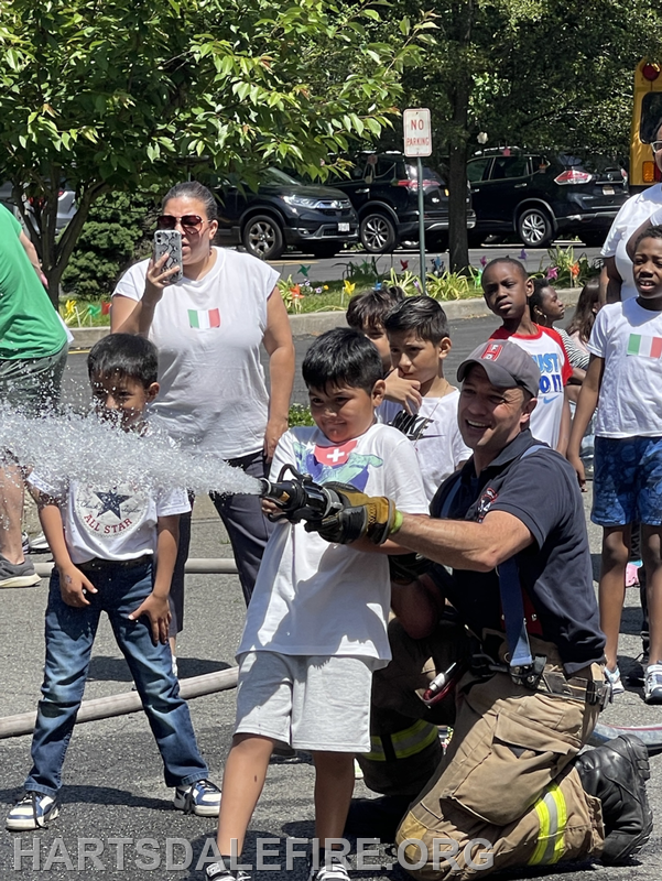 A firefighter helps a child spray water from a hose while a group of kids and an adult watches, enjoying a fun outdoor event.