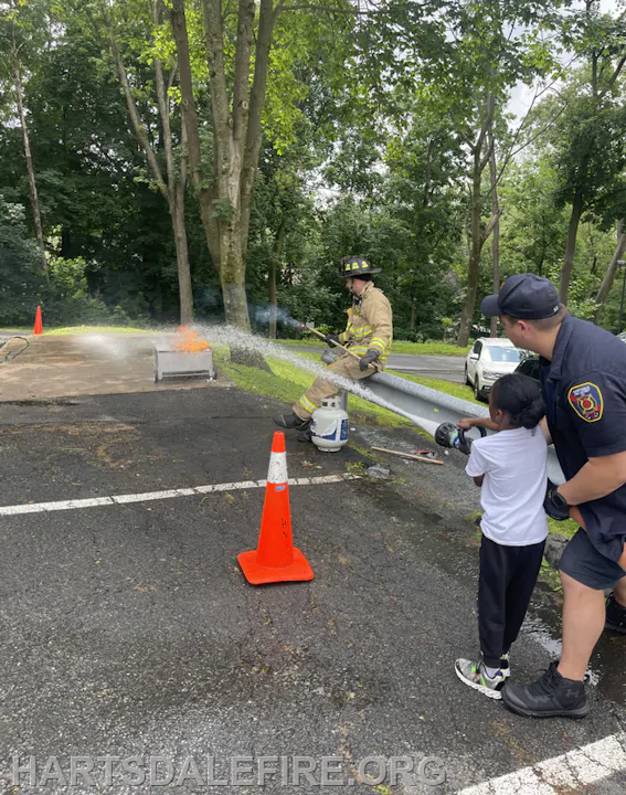 A firefighter and a child practice using a hose to extinguish a controlled fire, with safety cones in the area.