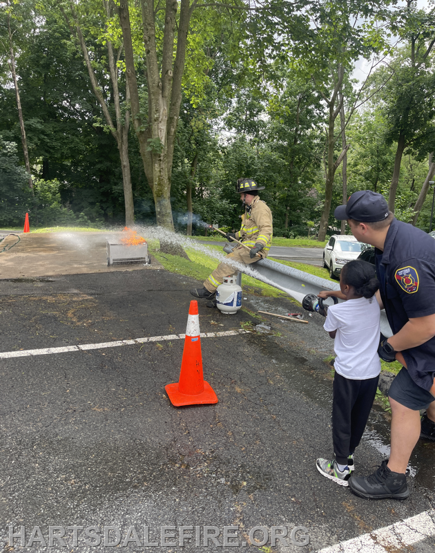 A firefighter and a child practice using a hose to extinguish a controlled fire, with safety cones in the area.