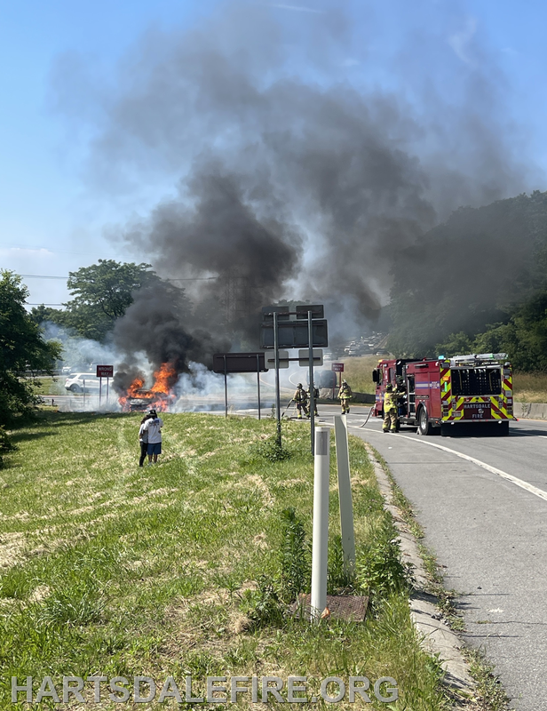 A vehicle is on fire, emitting thick smoke, while firefighters respond to the scene on the roadside.