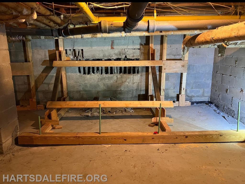 A basement with exposed pipes and wooden supports reinforcing a damaged wall.