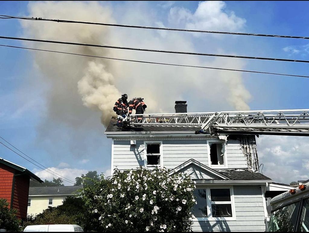 Firefighters are on a rooftop battling smoke from a house fire, with visible damage to the building and nearby plants.