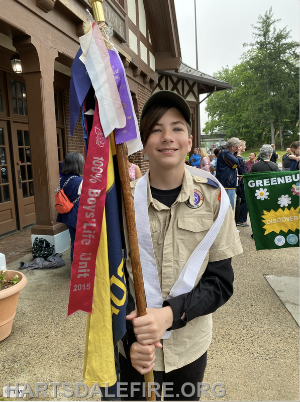 A smiling youth in a beige scout uniform holds a flagpole with colorful banners, standing outside a building among a crowd.