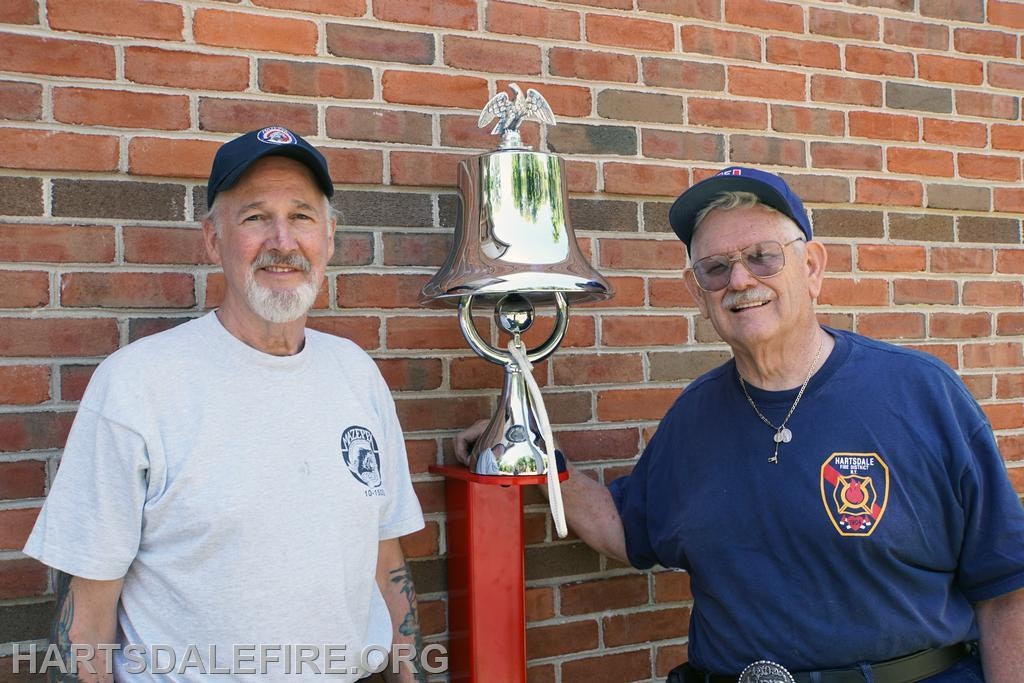 Two men stand next to a shiny bell on a red stand, against a brick wall, likely celebrating a fire department milestone.